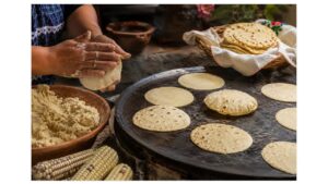 Mexican cook shapes fresh masa by hand while corn tortillas cook and puff on a traditional comal beside colorful ears of maize.