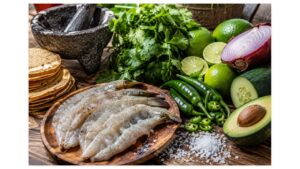 Fresh ingredients for authentic green aguachile including butterflied shrimp, serrano peppers, fresh limes, cilantro, cucumber, red onion, avocado, sea salt, tostadas, and a traditional Mexican molcajete.
