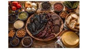 Traditional mole poblano ingredients including dried ancho, mulato, and pasilla chiles, Mexican chocolate, cinnamon, sesame seeds, almonds, pumpkin seeds, tortillas, bread, garlic, onions, and tomatoes arranged on a rustic wooden table.