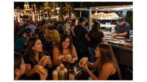 People enjoying authentic Sonoran hot dogs at a lively evening street food plaza in Hermosillo, Sonora, as a busy vendor prepares bacon-wrapped hot dogs under glowing string lights.