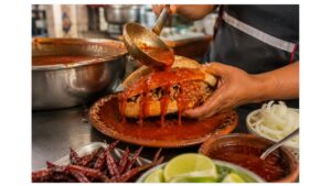 Artisan ladles tomato sauce over a carnitas-filled birote salado while preparing an authentic torta ahogada in Guadalajara.