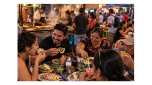 Mexican friends and family share tacos, tostadas, elote, and other street foods at a busy neighborhood food stand while cooks work nearby.